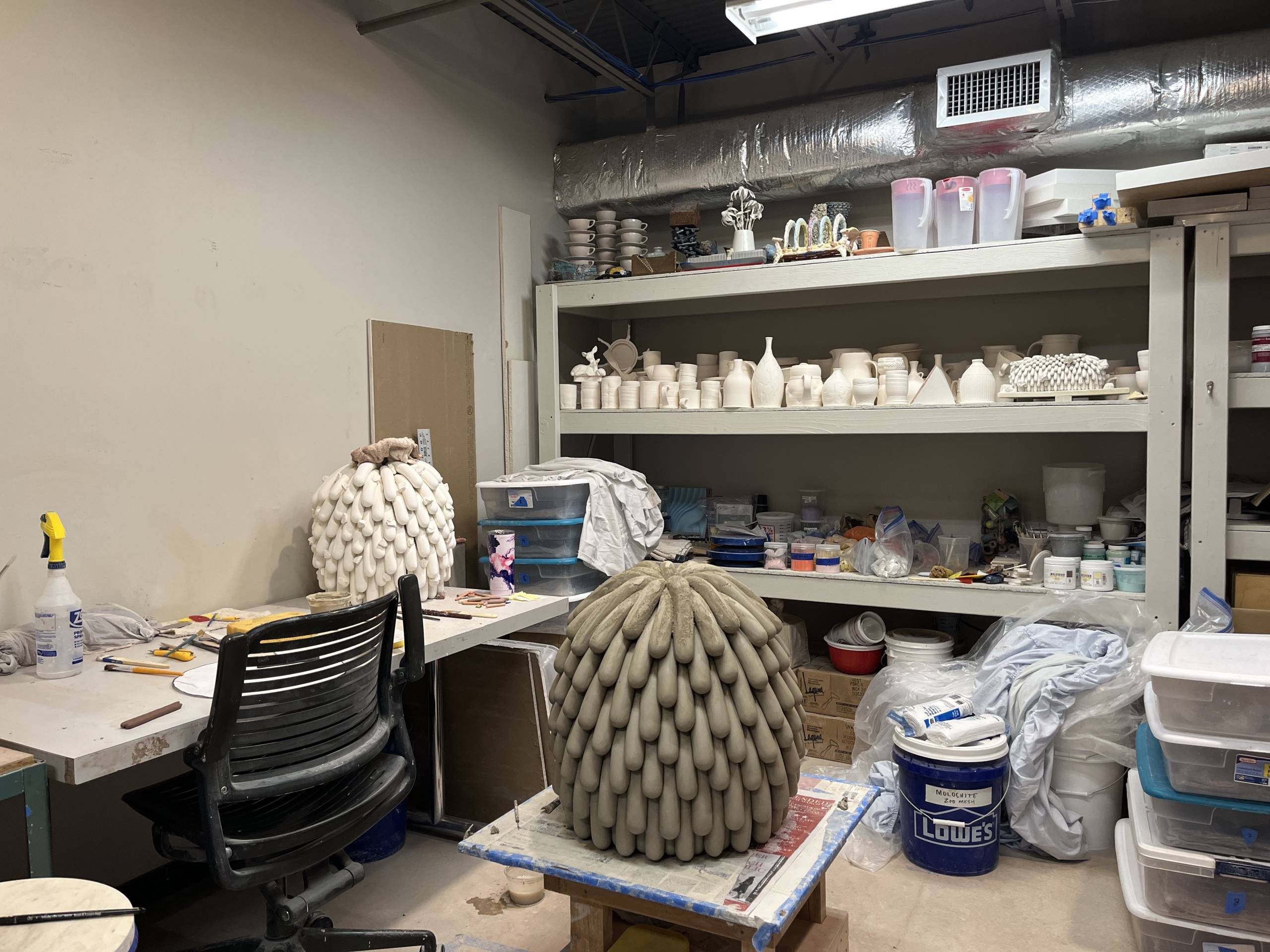 an artist studio with a gray clay work in progress on a workbench in the center, a work desk to the left, and shelves filled with white clay jars and art supplies in the background