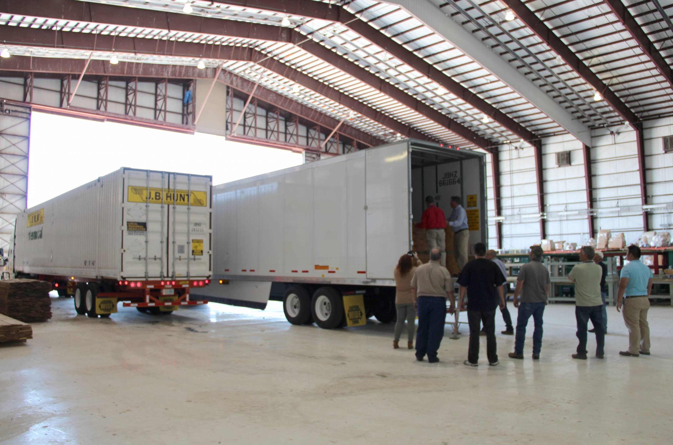 People in warehouse observe loading of J.B. Hunt trailer, metal beams and natural light visible.