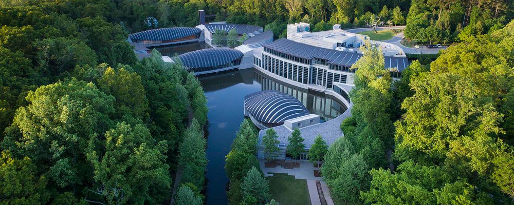 Modern architecture with curved roofs amid dense forest by a reflective lake.