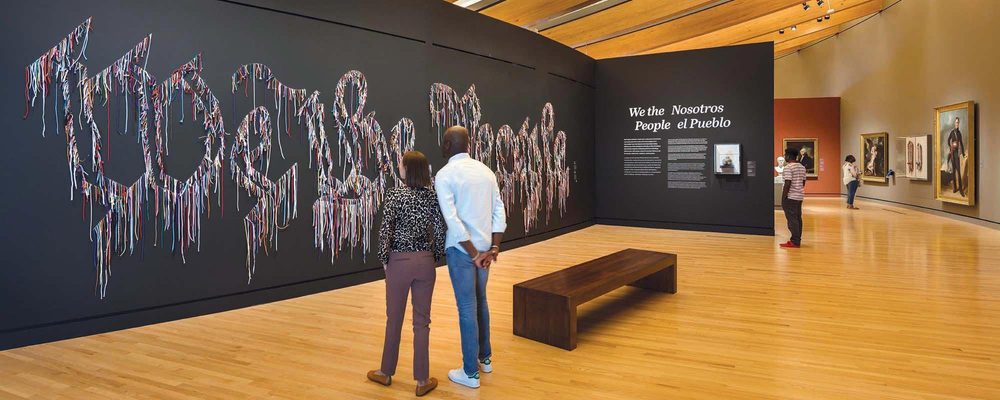 Galería de arte con visitantes, texto con cordones multicolores, cuadros enmarcados y paneles bilingües.