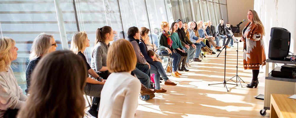 Woman speaking into a microphone, audience seated in bright room with large windows.