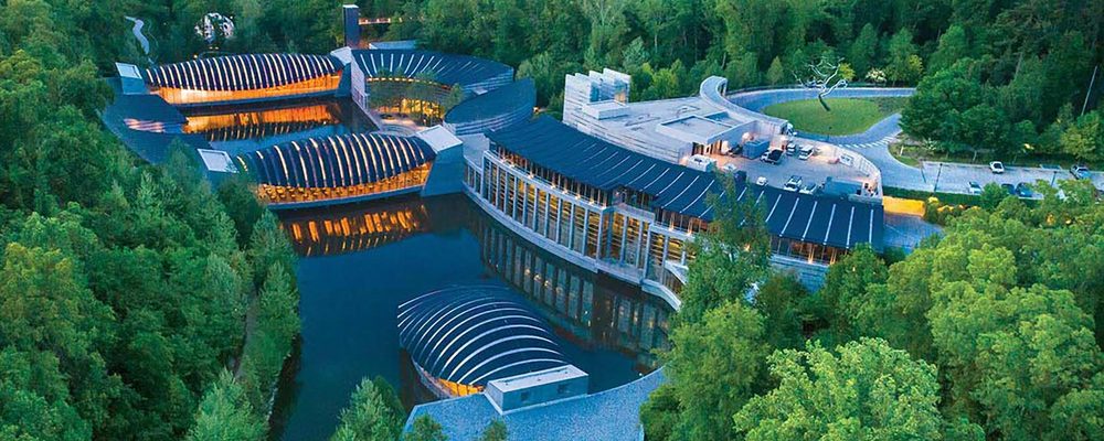 Modern complex with curved roofs and interconnected buildings over water, surrounded by forest.