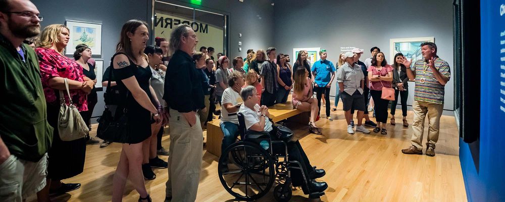 Group in museum listens to speaker; diverse audience, artworks on walls, includes wheelchair user.