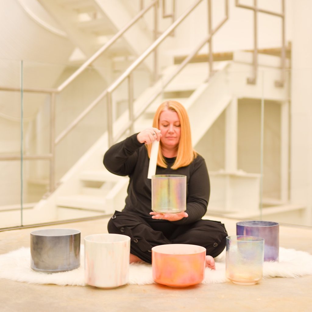 Woman sitting on white rug playing six crystal singing bowls in modern interior.
