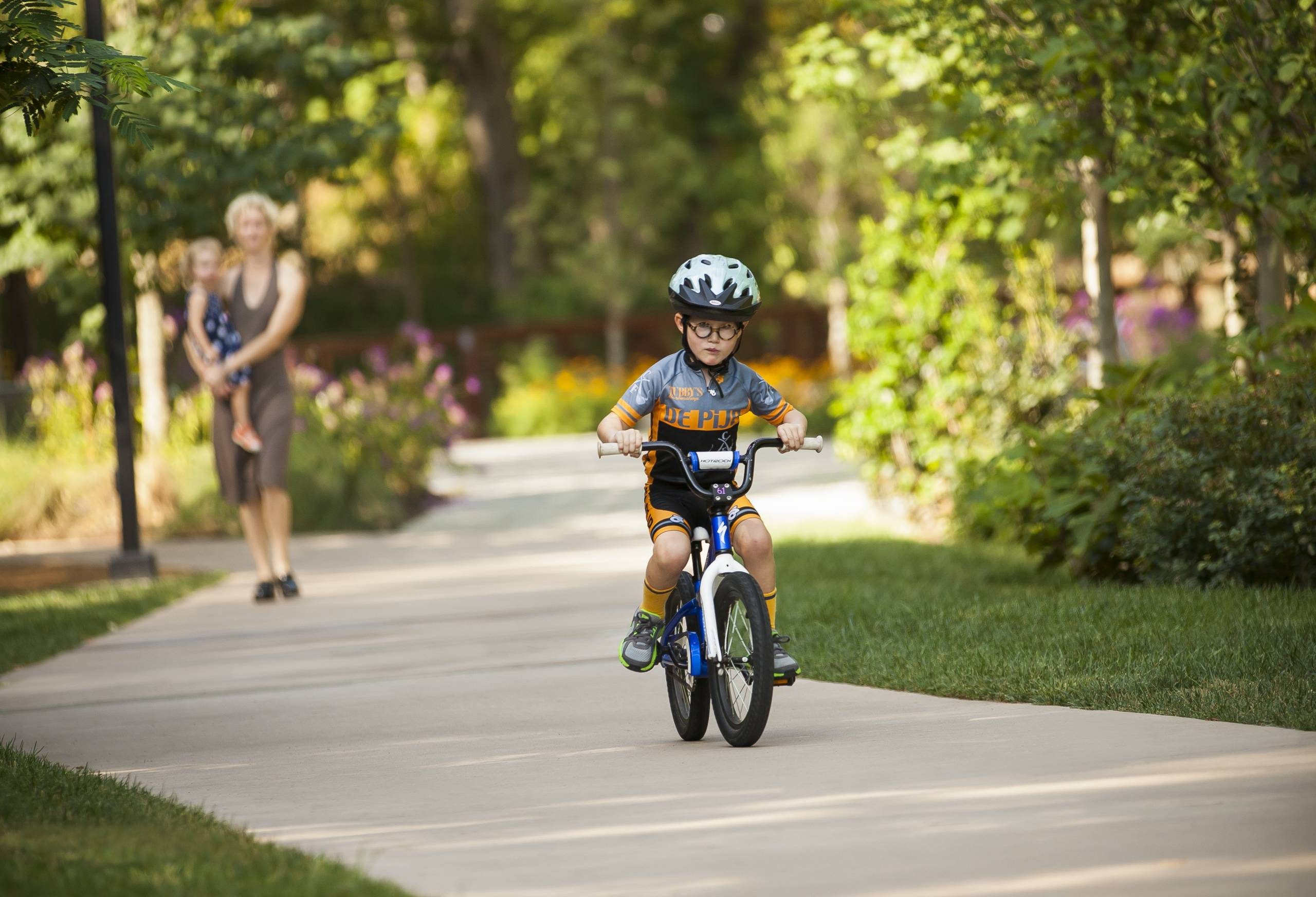 Child on bicycle with helmet rides on park path, adult with toddler in background, green trees.