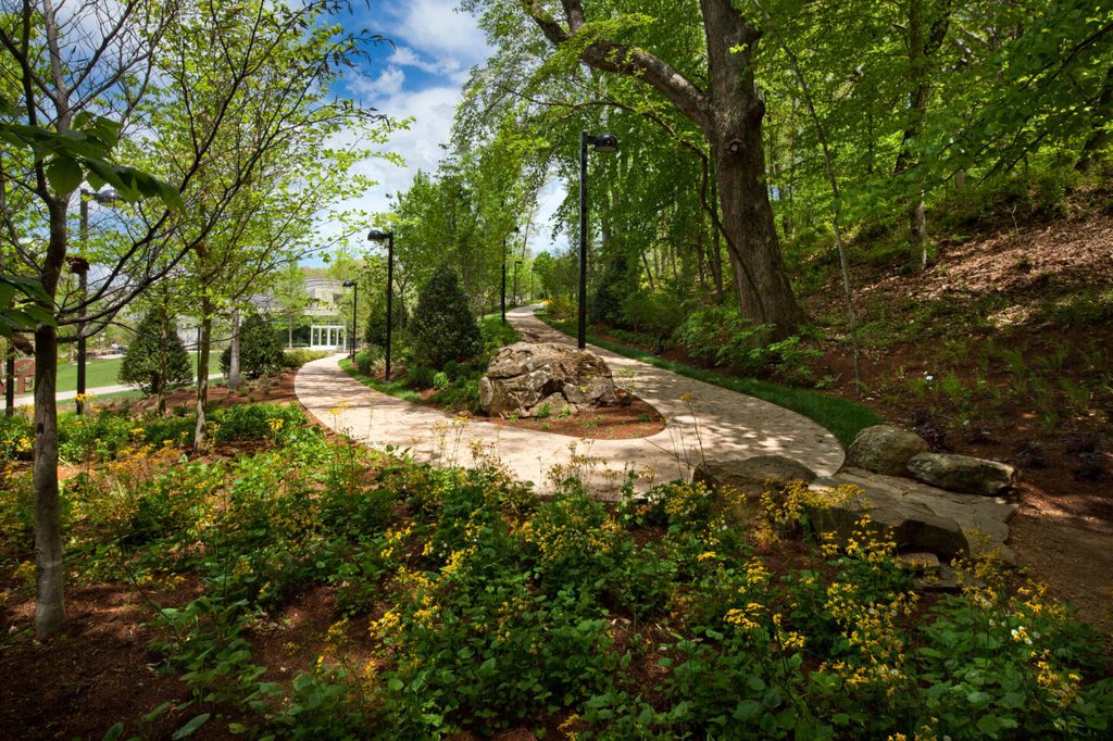 Scenic garden path with trees, flowering plants, large rock, and black lamp posts.