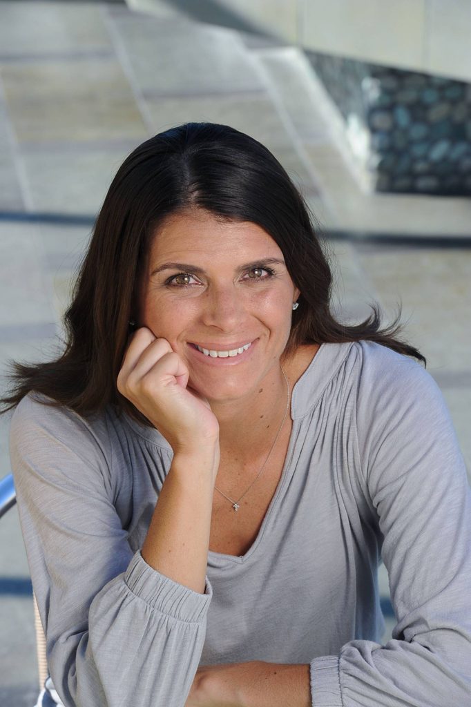 Woman with long dark hair wearing a light gray top and cross necklace sitting outdoors and smiling.