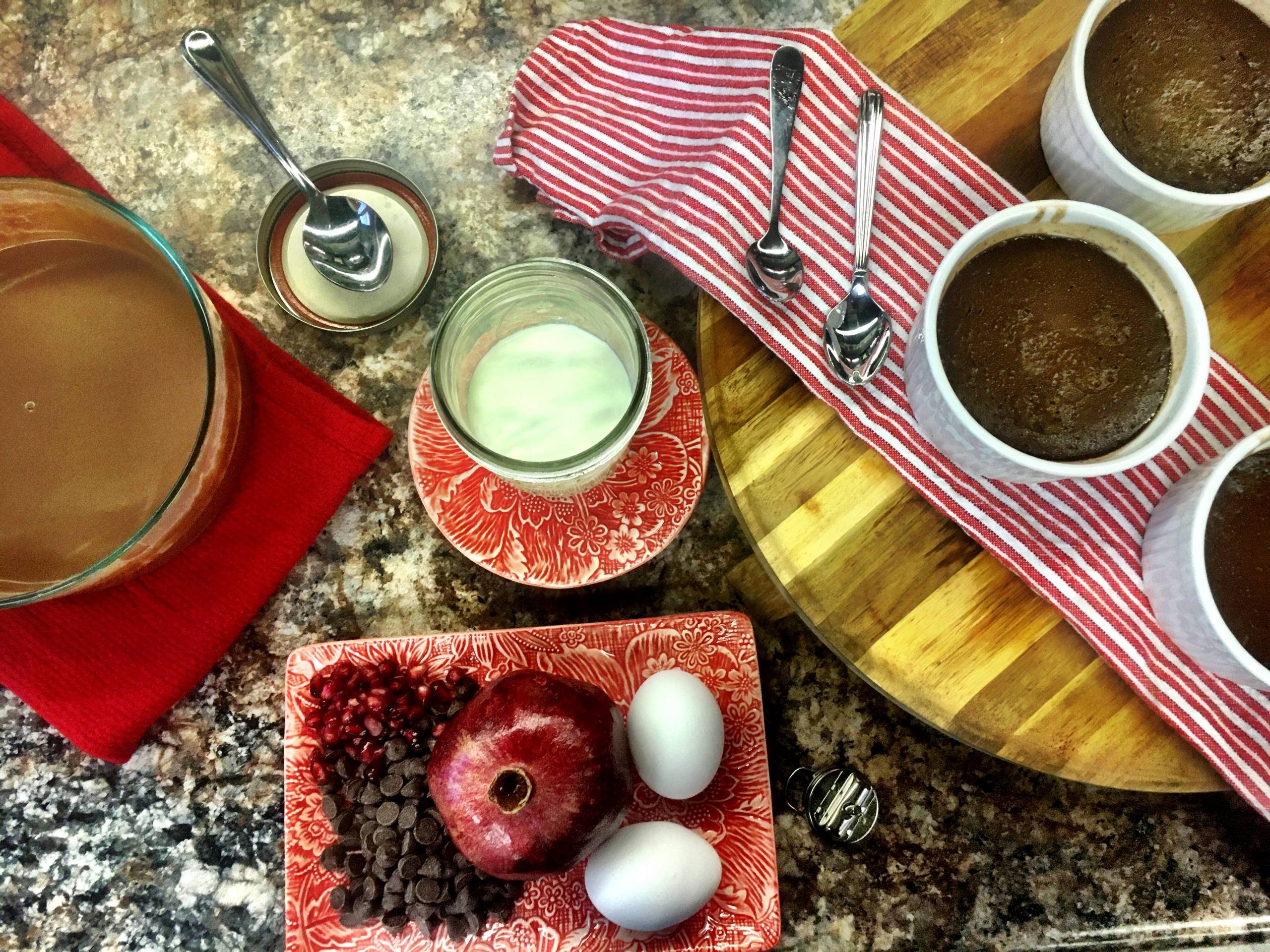 Tabletop with chocolate ramekins, pomegranate, eggs, milk, chocolate chips, and red striped cloth.