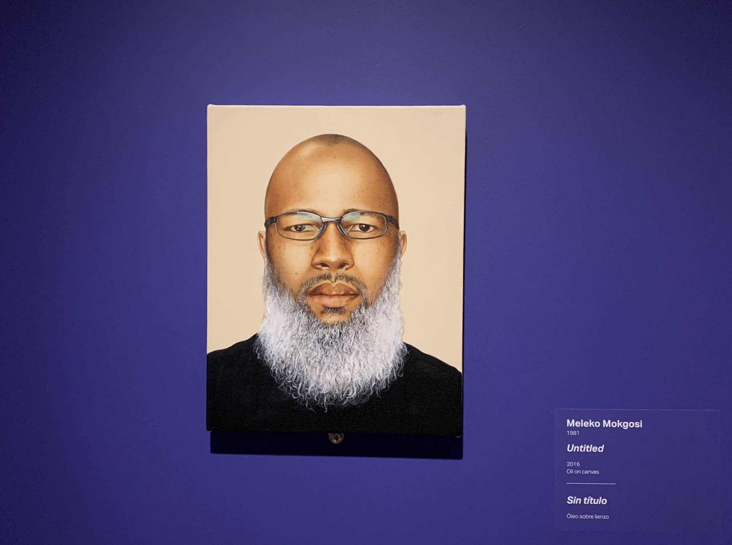 Self-portrait of man with grey beard wearing glasses hanging on wall with exhibition labels beside it in Crystal Bridges at 10