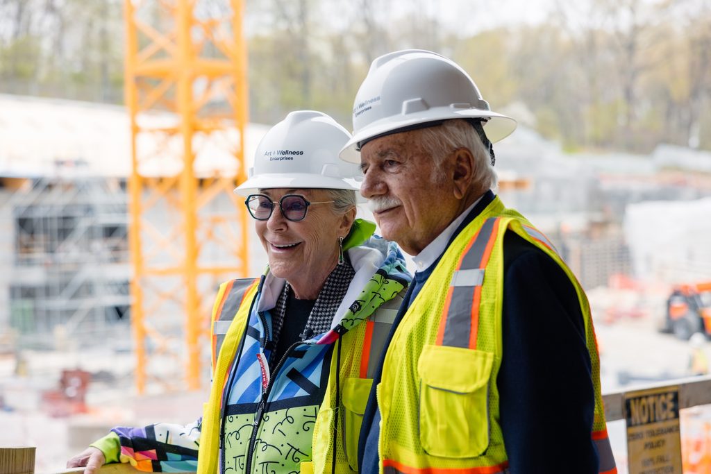 Two people in hard hats and vests at a construction site with scaffolding and crane visible.