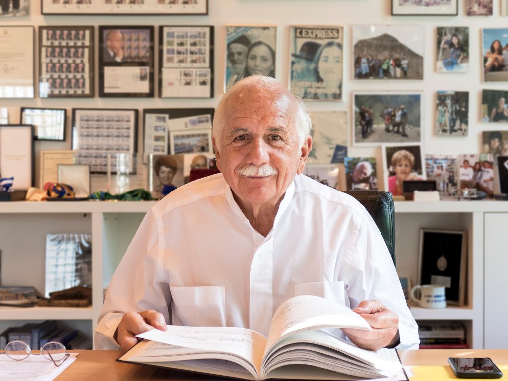 Elderly man in white shirt sitting at desk with open book, glasses, phone, and photo-filled wall behind him.