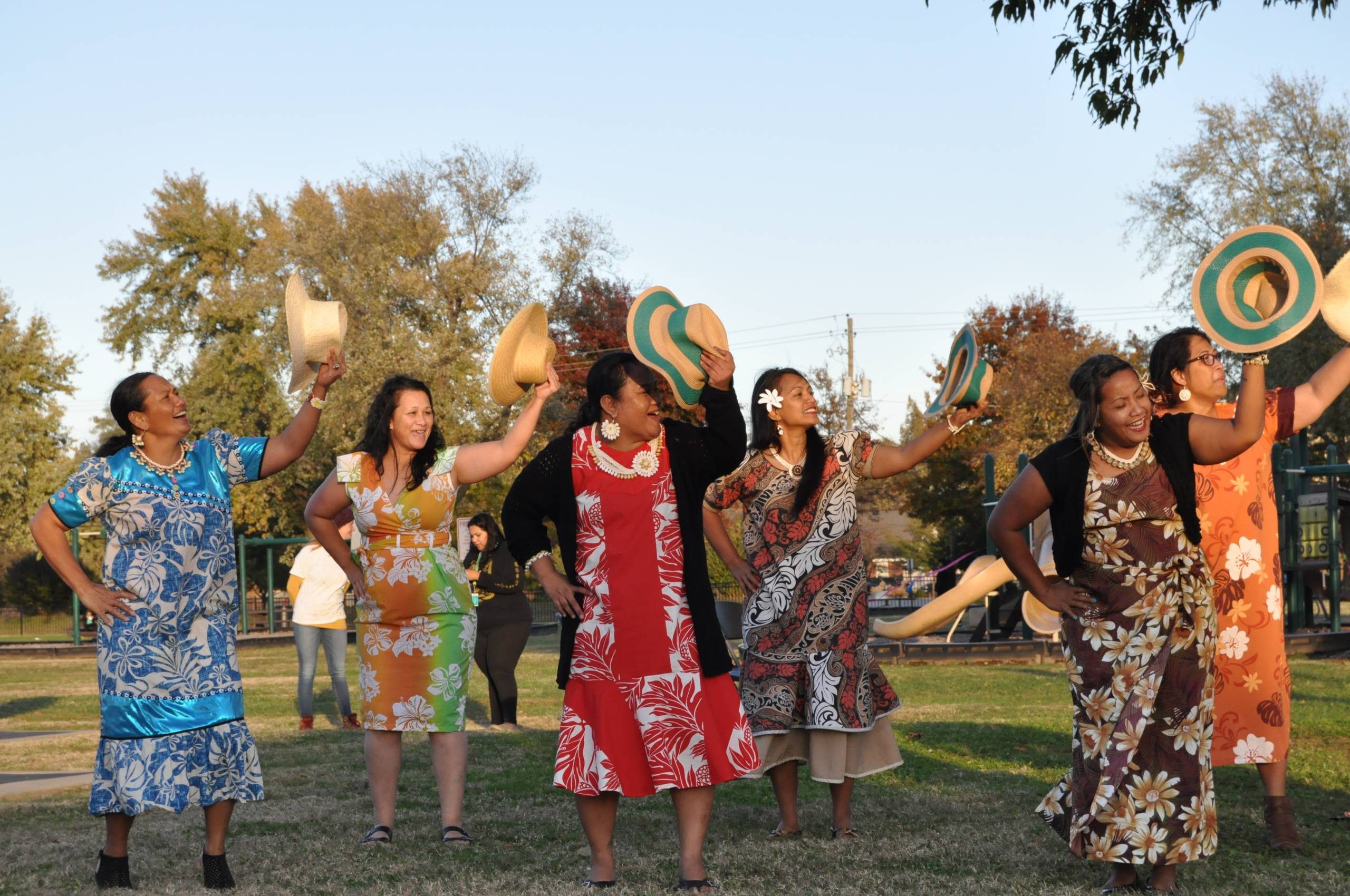 Women in colorful dresses performing a dance outdoors, holding straw hats on a sunny day.
