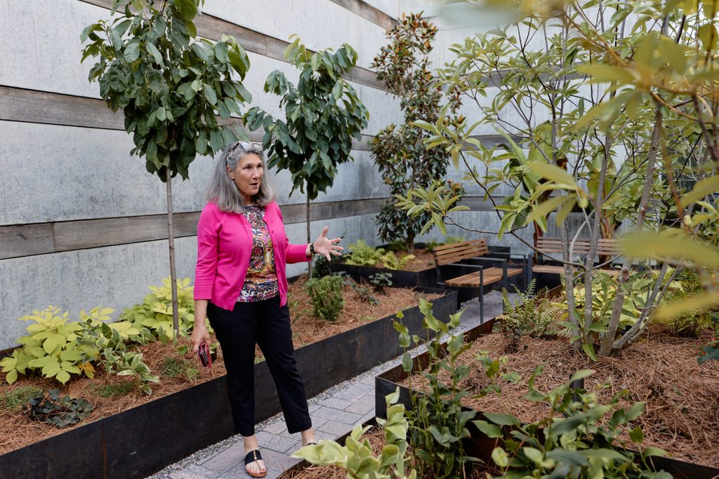 Woman in pink cardigan standing in an outdoor garden with raised beds and benches.