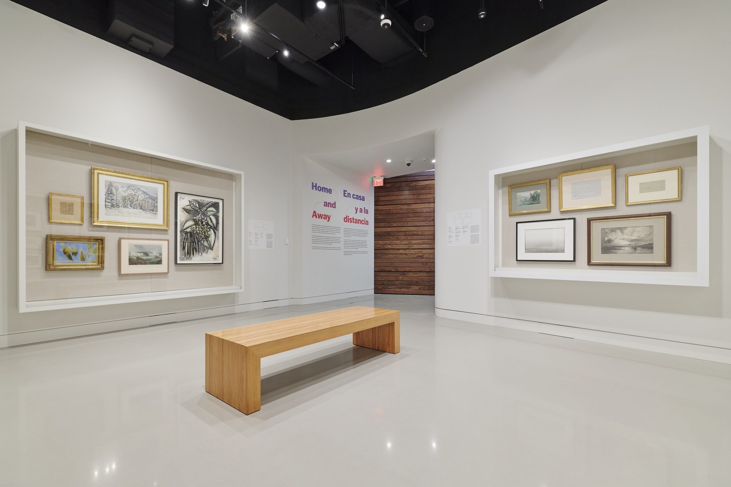 Modern art gallery room with framed art, wooden bench, bilingual text, and exhibit signage.