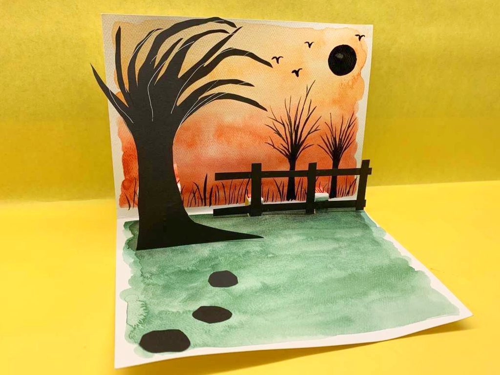 create an eerie pop-up book art activity; image shows a painted and decorated pop-up card featuring a tree, fence, and birds