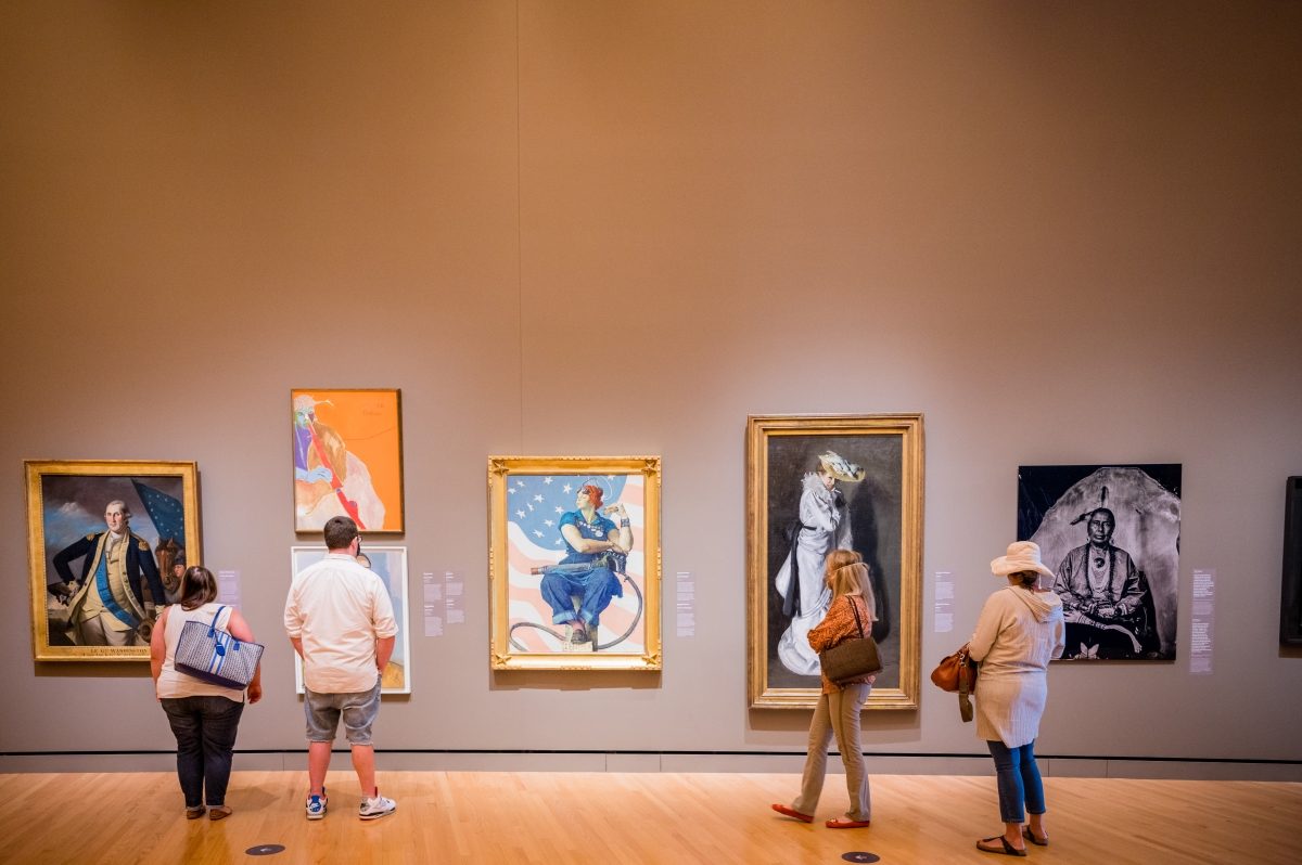 People viewing various framed artworks in a gallery with wooden floors and neutral walls.