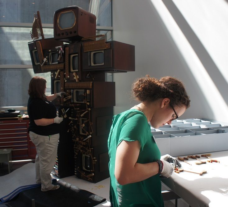 Two people assemble a structure of vintage TVs and wood indoors; one works on a table nearby.