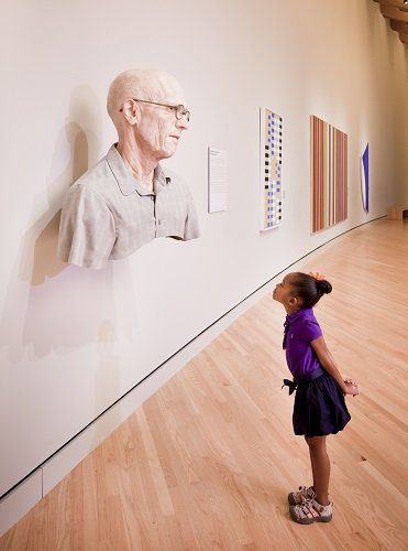 Young girl observes realistic elderly man's sculpture in art gallery.