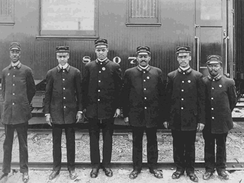 a row of black train conductors in uniform with caps and shined shoes standing in front of a train car