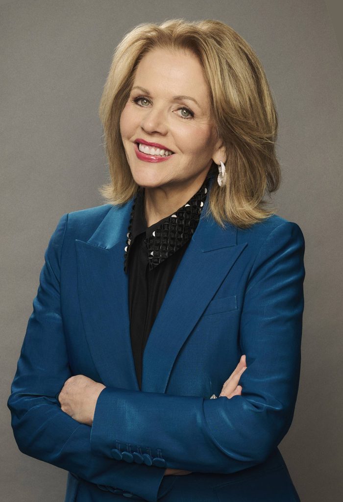 Blonde woman smiling with arms crossed wearing a blue blazer and black shirt on gray background