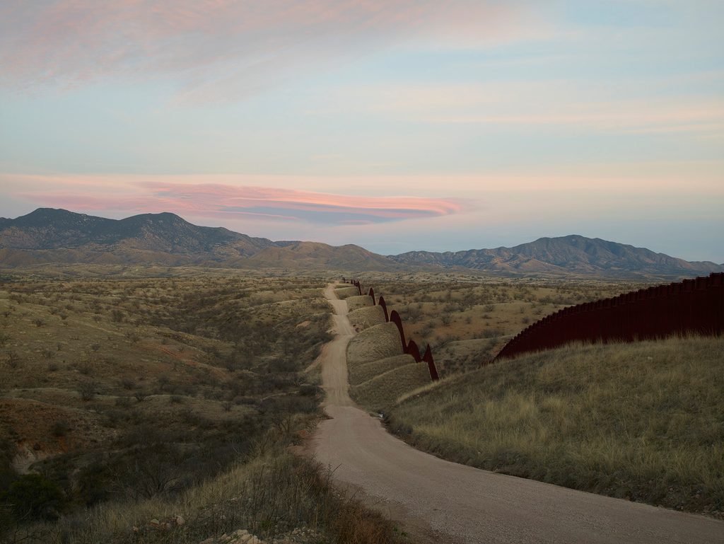 Rural landscape with border wall, dirt road, hills, and pastel sky at sunset.
