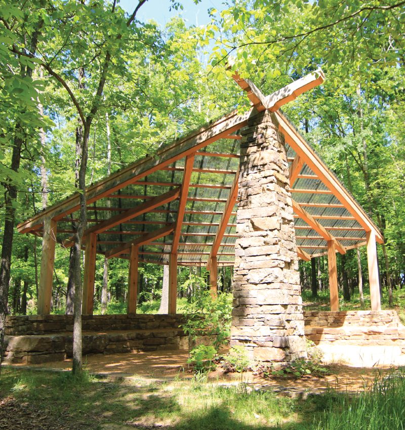 Wooden pavilion with stone pillar and transparent sloped roof in a lush forest setting.