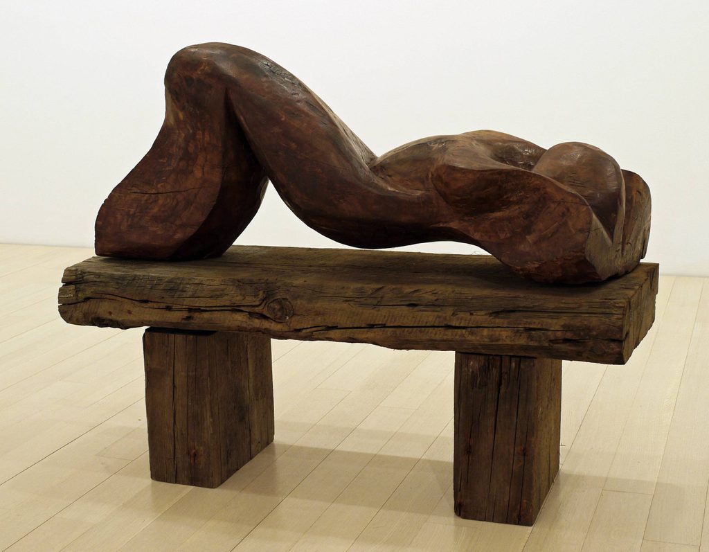 Abstract wooden sculpture of a reclining figure on a wooden base with block supports.