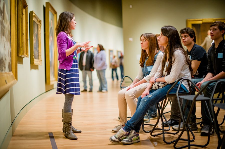 Woman speaking to seated young adults in an art gallery, surrounded by paintings.