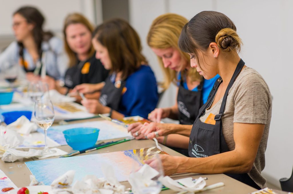 Individuals wearing aprons painting at a table with brushes and various art supplies.