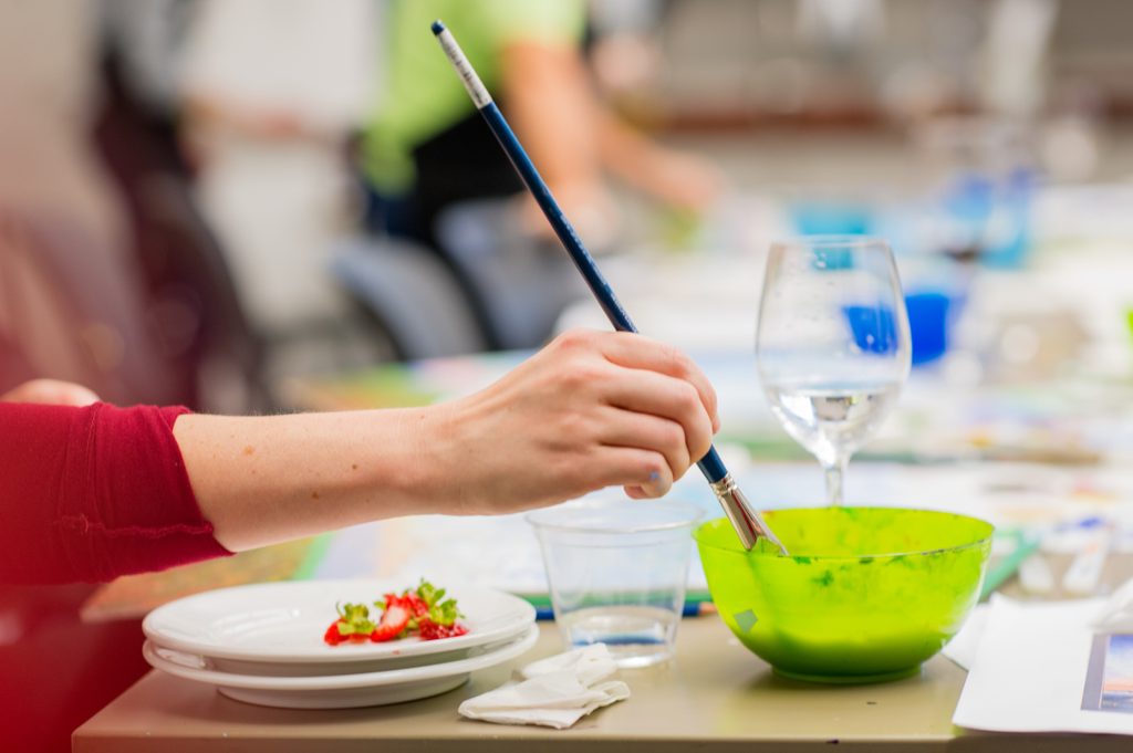 Hand holding paintbrush dipped in green bowl on table with plates and glassware.