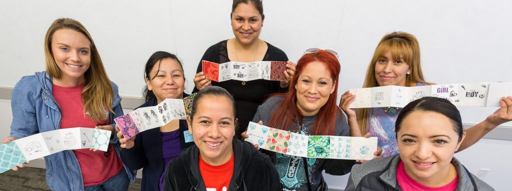 Six women smiling, holding accordion-style papers with various designs in an indoor setting.