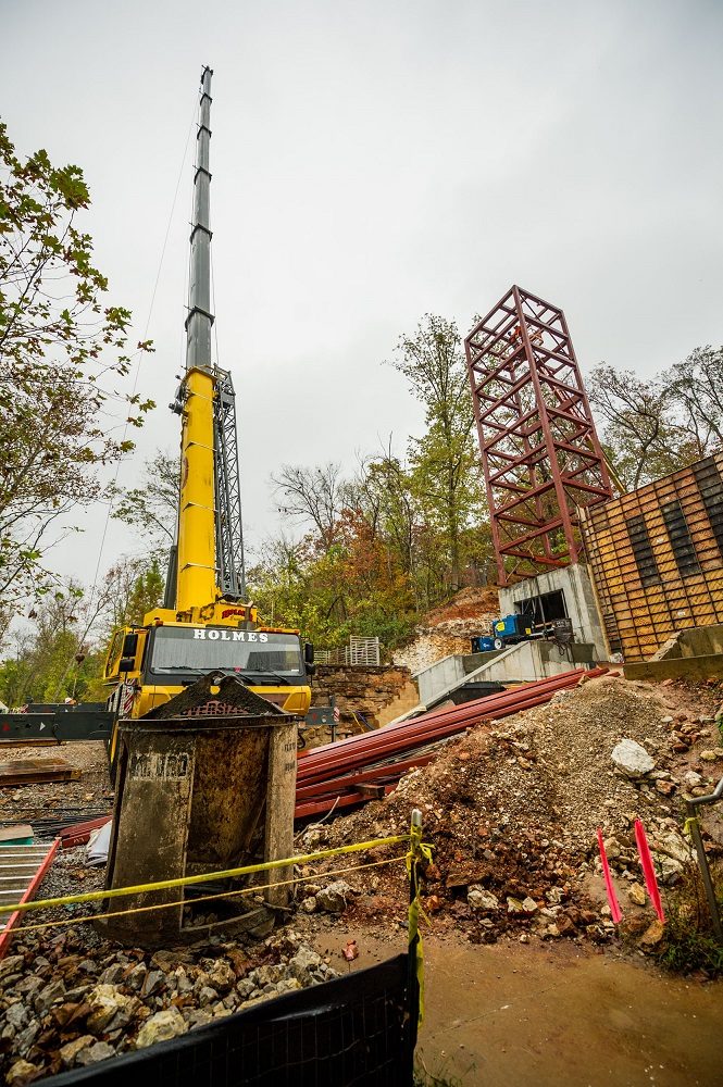 Large yellow crane next to red steel framework at a construction site amid wooded surroundings.