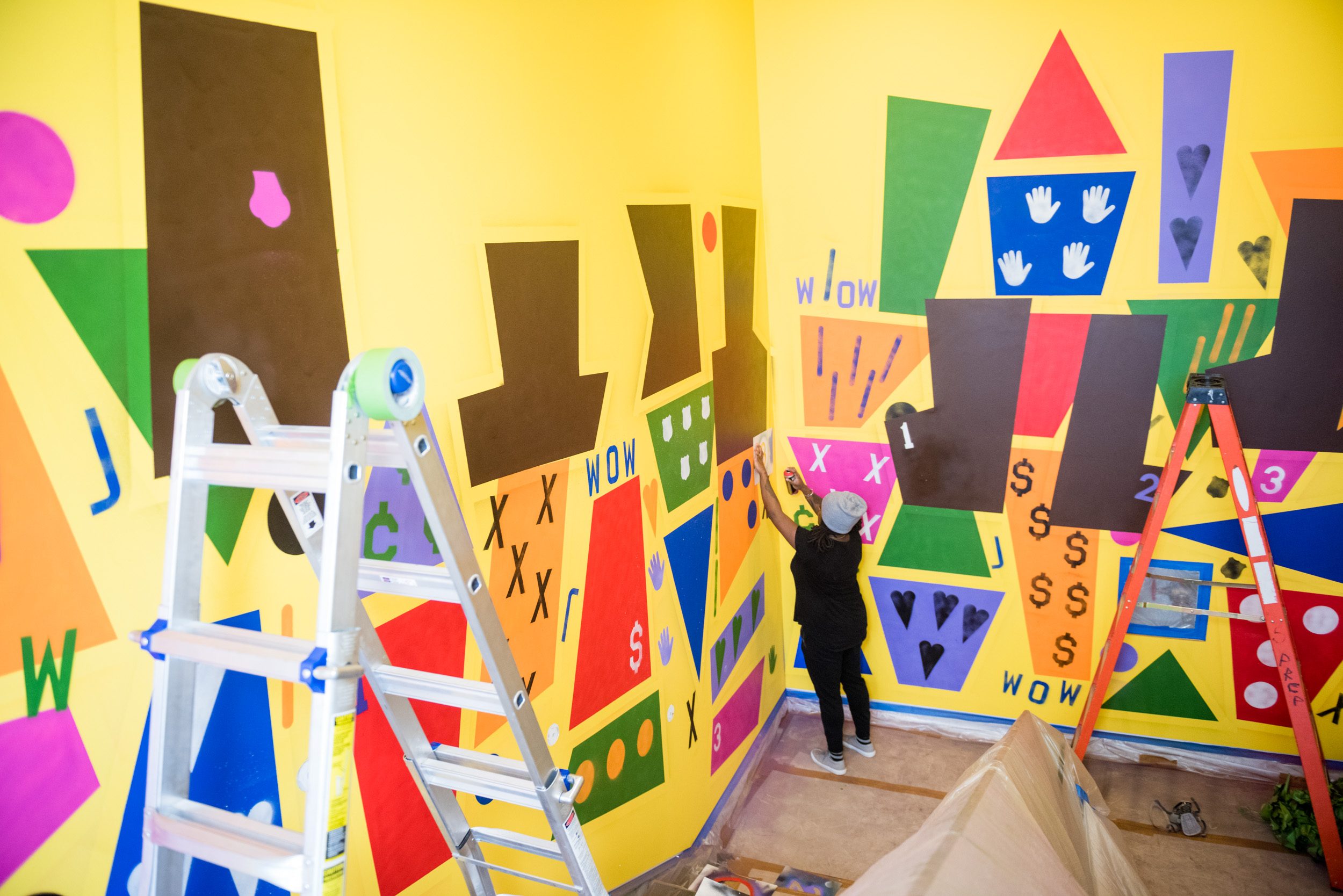 Person painting colorful mural on yellow wall with shapes, letters, and numbers; ladders nearby.