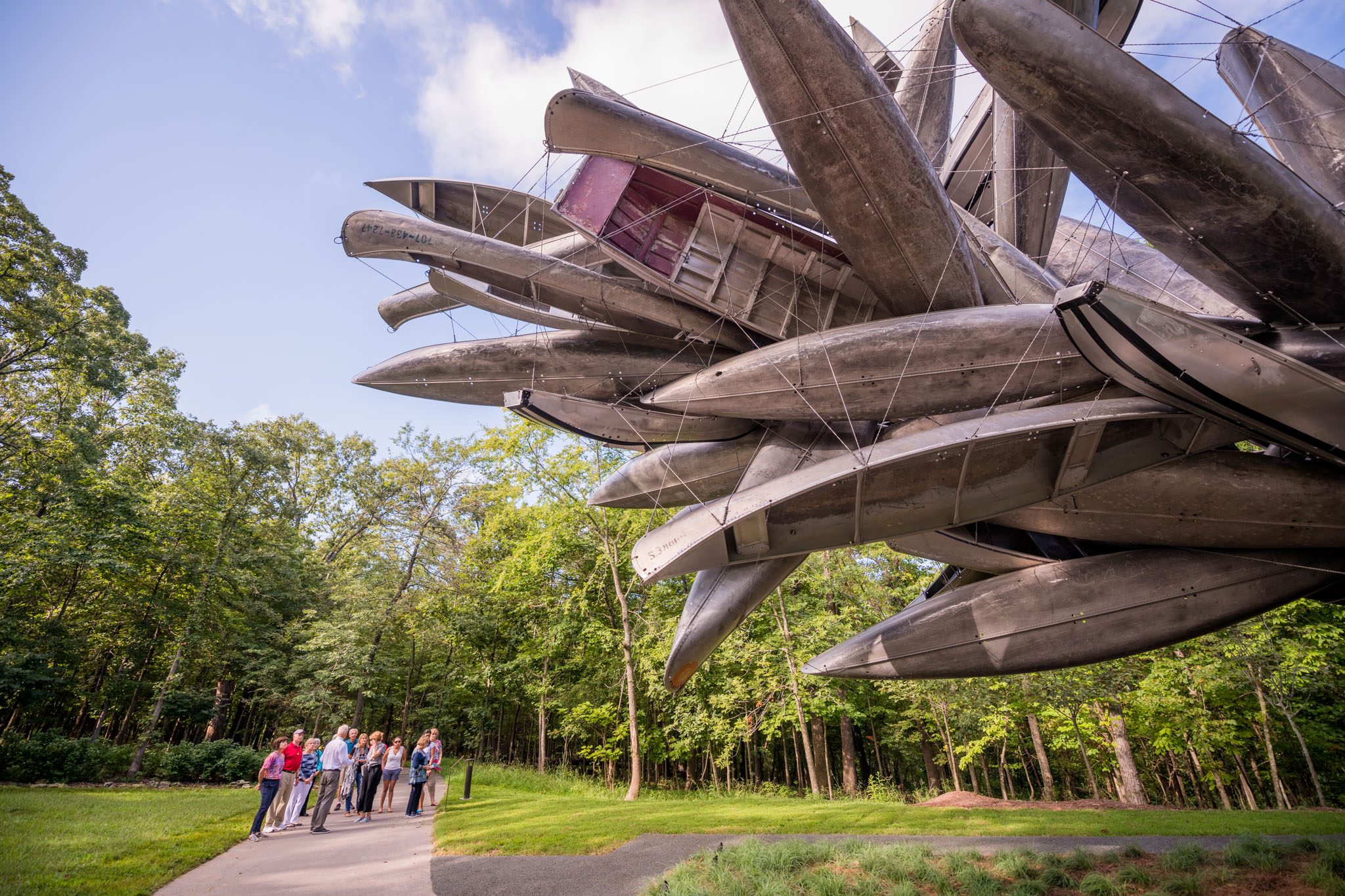Group observing large sculpture of canoes suspended among trees in wooded area.