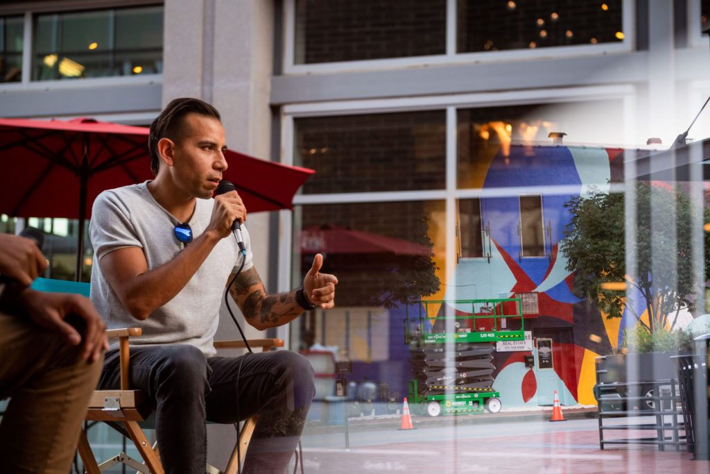 Person speaking into a microphone outdoors with red umbrella, mural, windows, and trees خلفية.