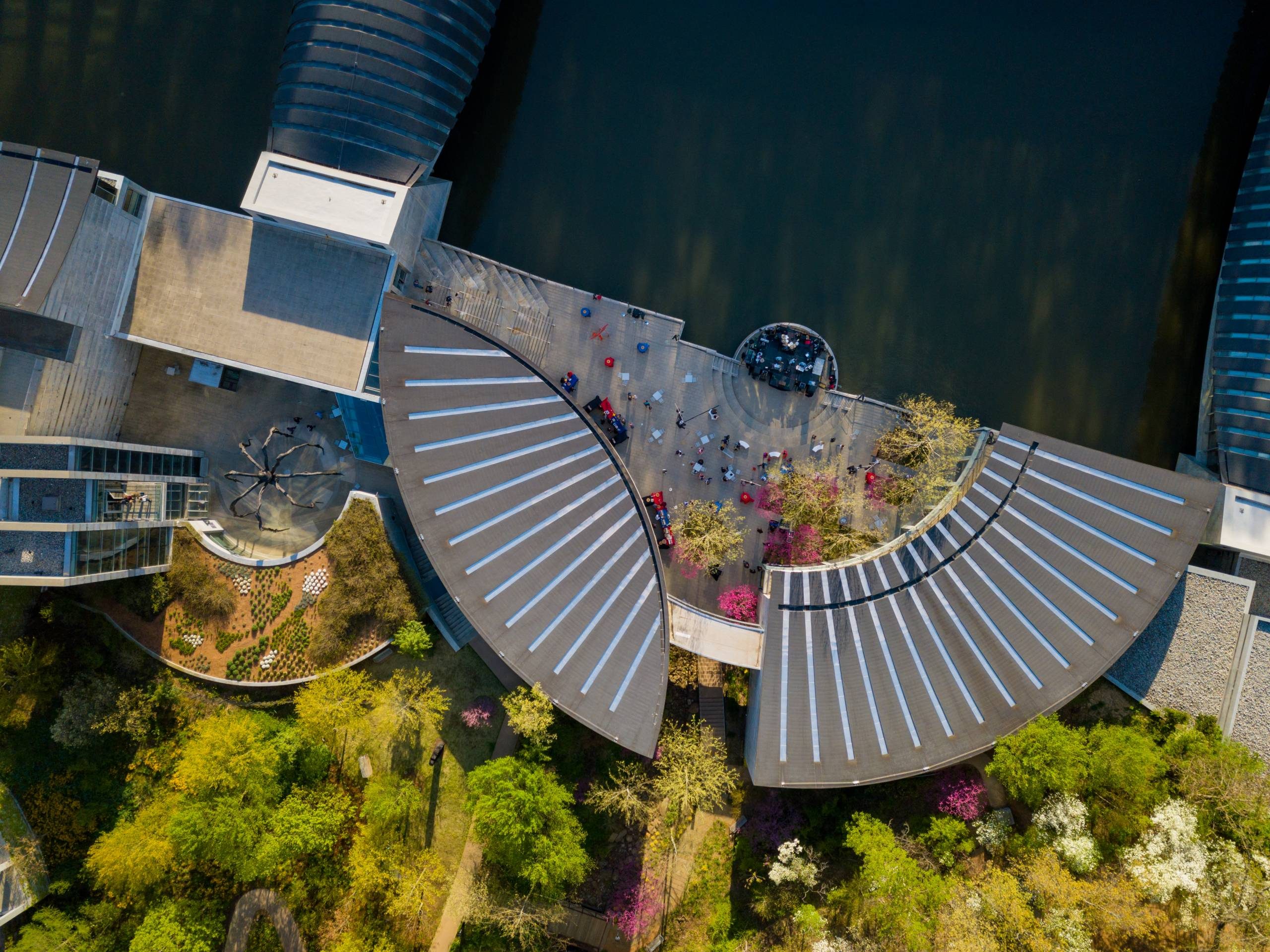 Aerial view of modern complex with curved roofs, gardens, and seating near water.