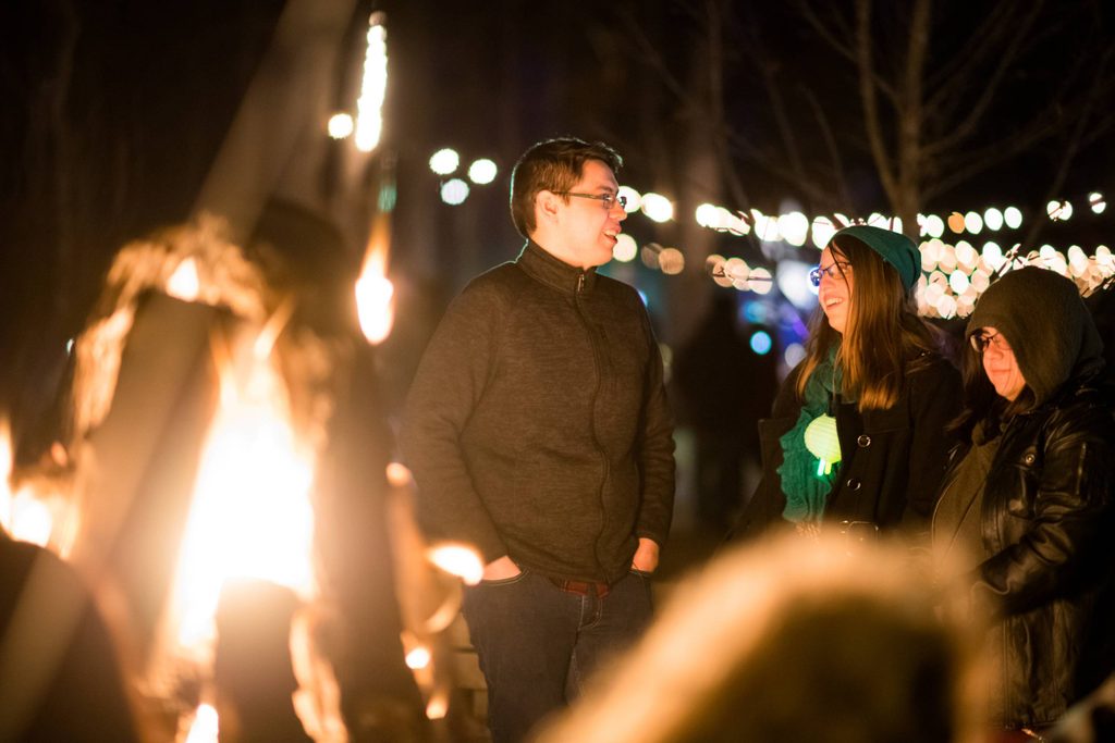 Group gathered around a fire at night, wearing warm clothing, with blurred decorative lights.