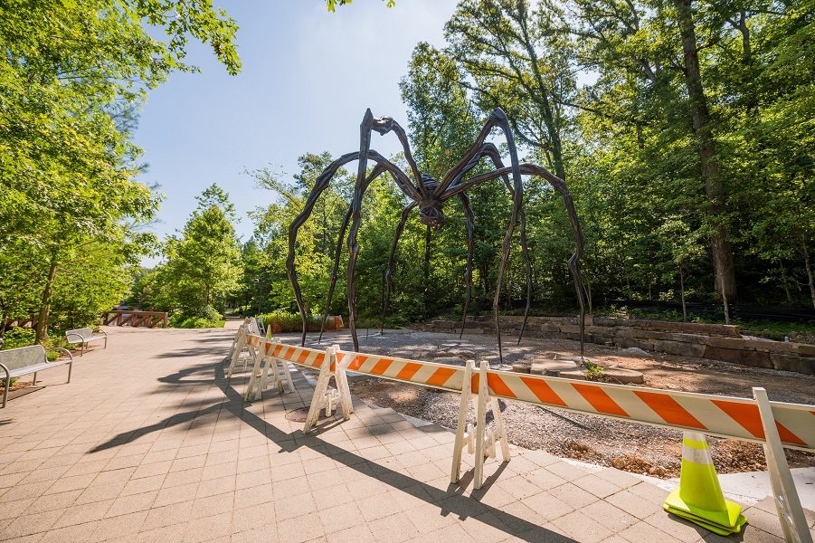 Large outdoor spider sculpture in wooded area, surrounded by barricades on a paved path.