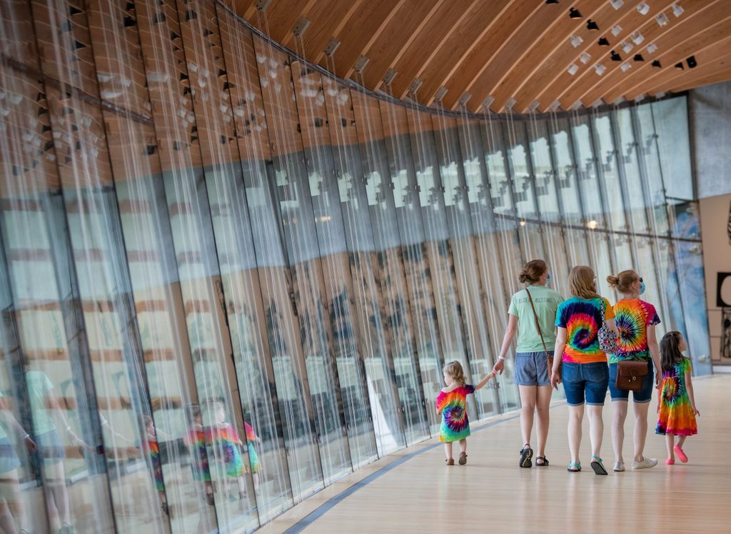 People in tie-dye clothes walking in a glass-walled hallway with a wooden ceiling.