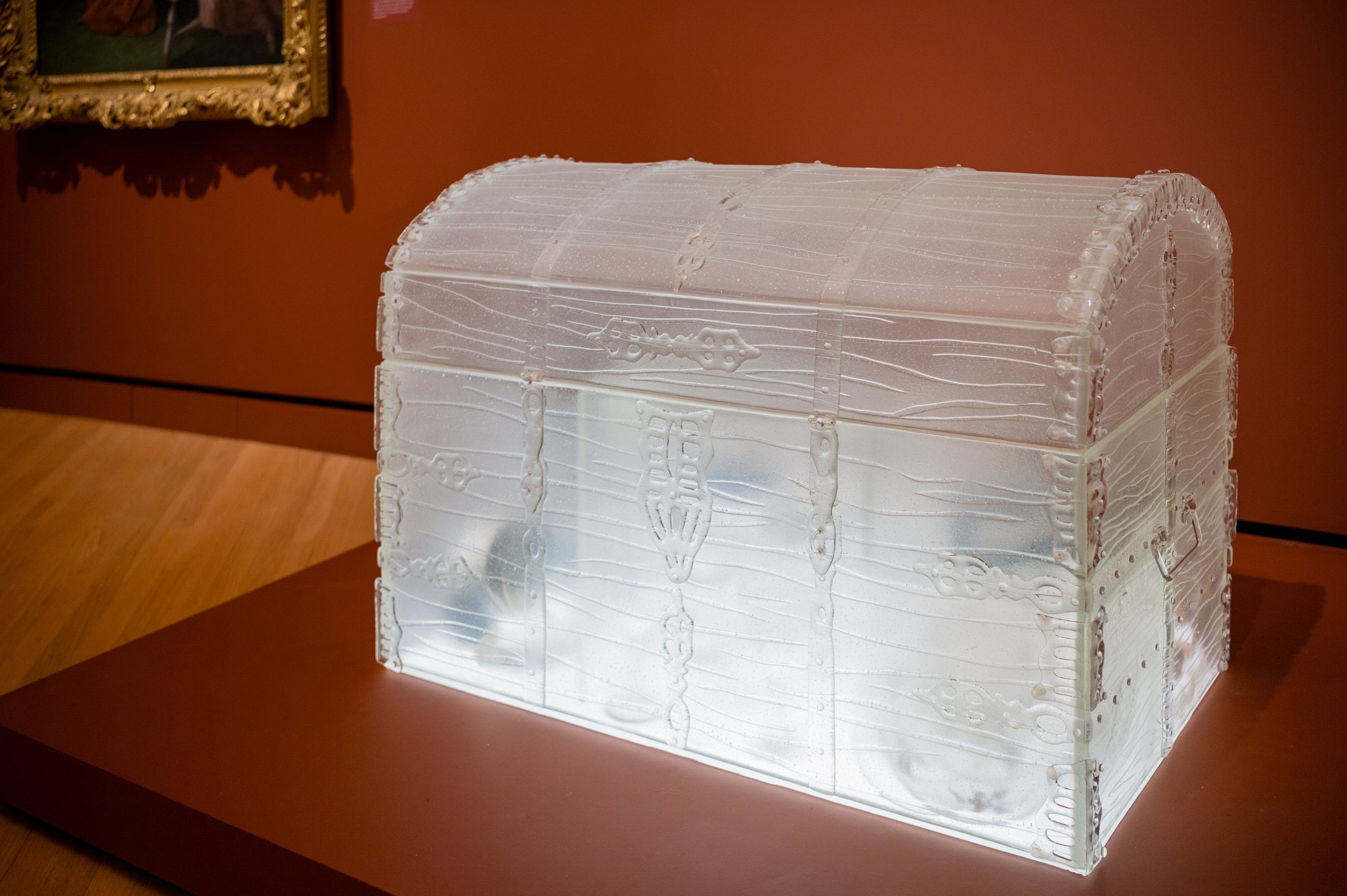 Transparent glass treasure chest with intricate designs in a museum, painting in background.