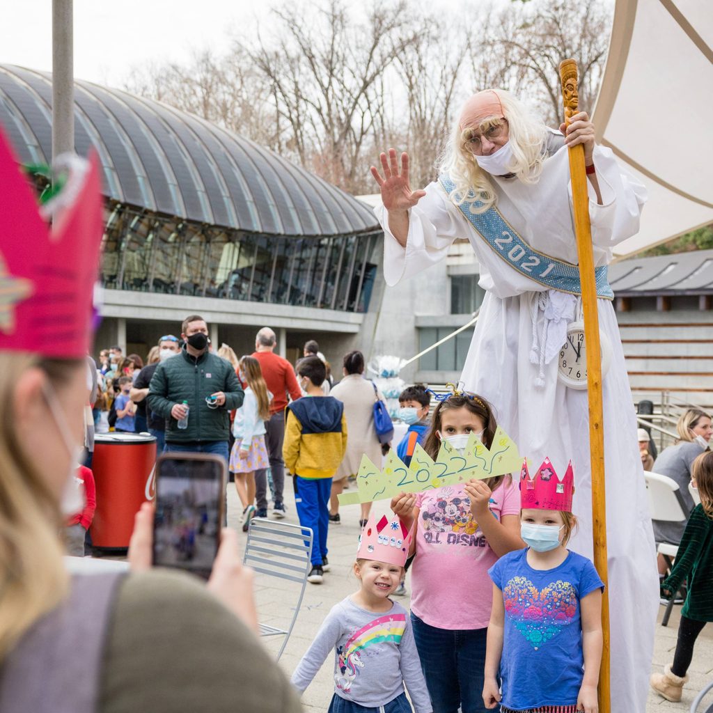 a man dressed as father time in a wig, walking on stilts, and carrying a cane poses for a photo with three children wearing paper crowns