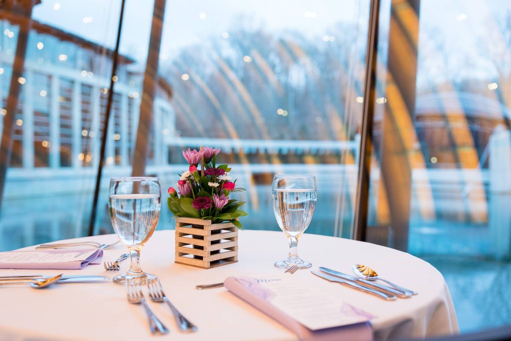 A tableclothed table setting for two with drink glasses and centerpiece flowers looking out onto the Crystal Bridges upper pond in winter
