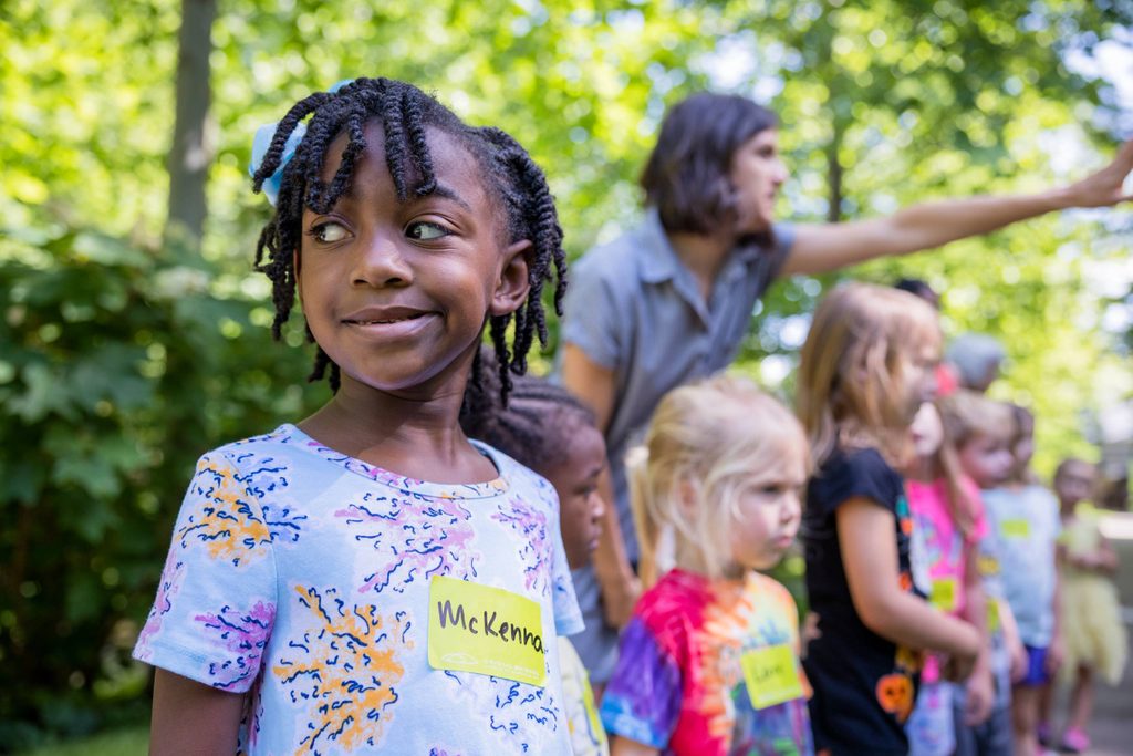 A group of children is outdoors, with name tags on their shirts. A child in the foreground is smiling, and another adult in the background appears to be guiding or instructing them. The setting is a lush, green park or garden.