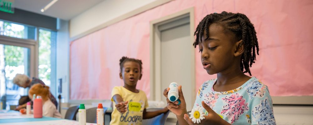 Children crafting in a bright room with glue, daisy, and colorful bottles on the table.