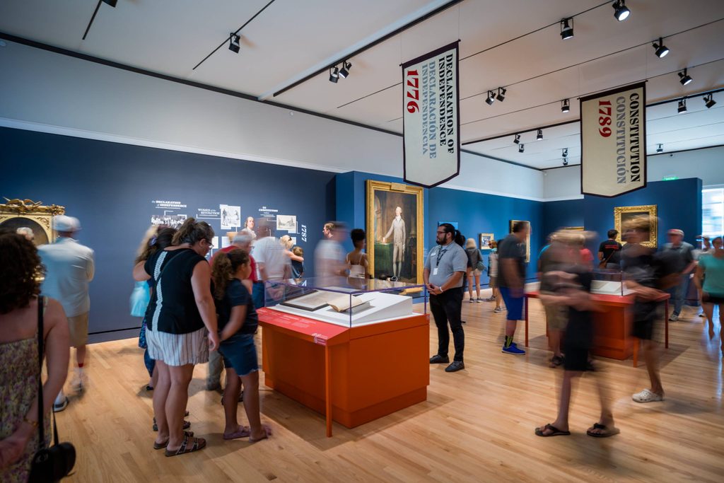 a group of people look at documents encased in glass on a pedestal and walk around a gallery space