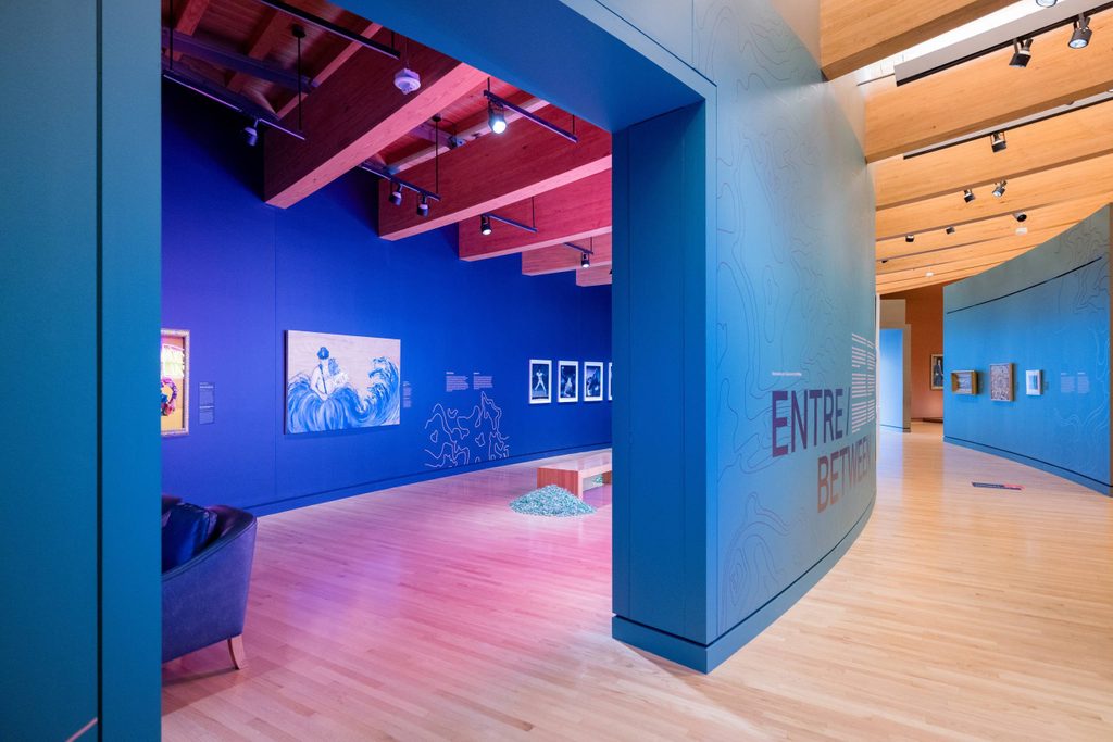 a gallery view of an ocean blue wall and entrance opening to a brightly lit and colorful gallery with works on the walls and in the space