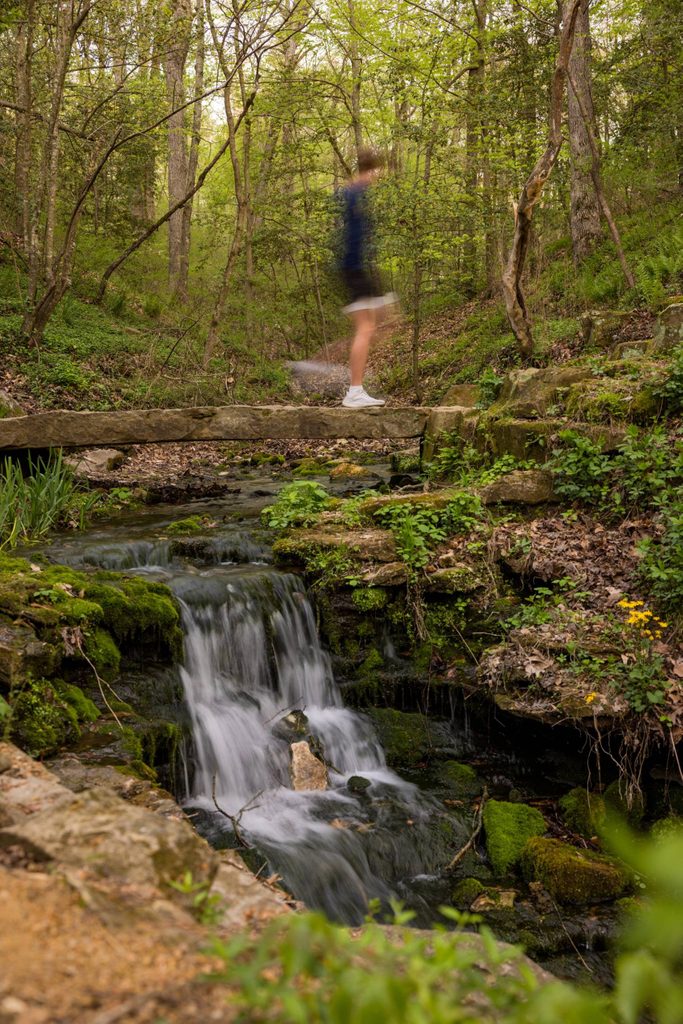 Una persona aparece borrosa cruzando un estrecho puente de piedra sobre una pequeña cascada en un frondoso bosque verde.