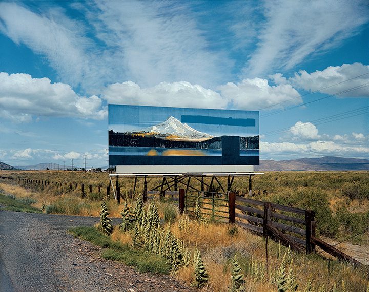 Roadside billboard shows snowy mountain, sky, grass, and fence in rural landscape.