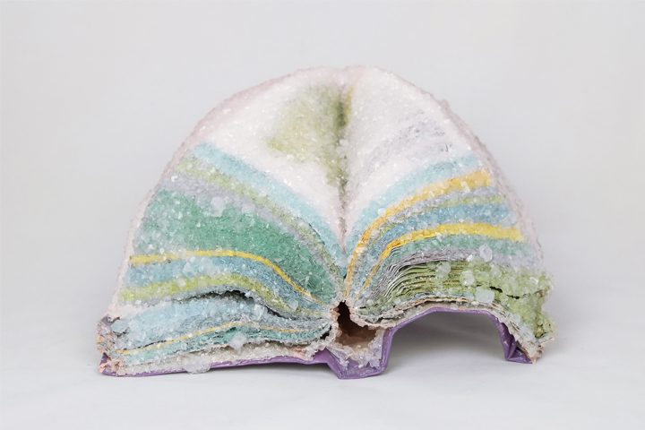 Sculpture of open book with crystal-like texture, layered in green, blue, yellow, and purple.