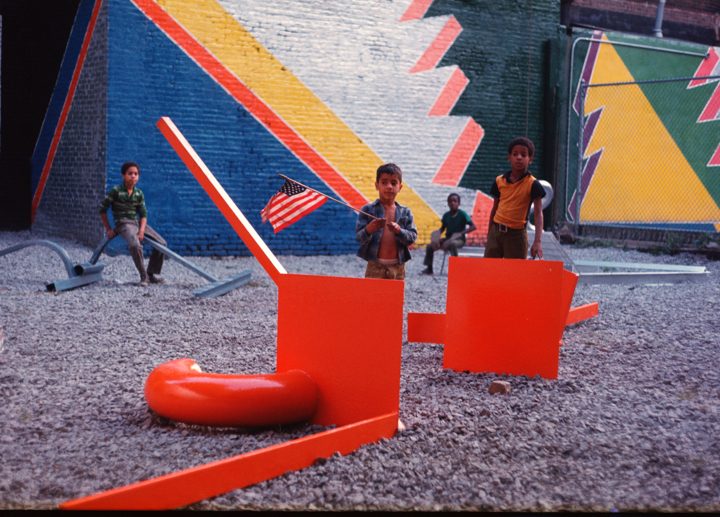Children play in a gravel area with red structures and a colorful geometric mural in the background.